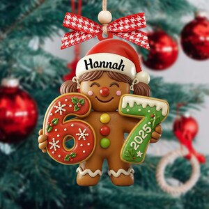 May include: A gingerbread-style Christmas ornament featuring a smiling girl wearing a Santa hat with the name "Hannah". The ornament holds the numbers "6" and "7" with "2025" on the "7". It is decorated with red and white accents.