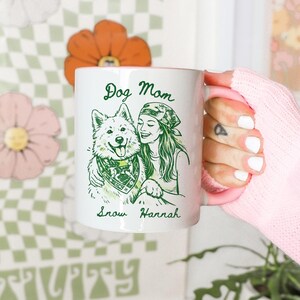 Może przedstawiać: Biały ceramiczny kubek z r&oacute;żowym wnętrzem i uchwytem, ozdobiony zieloną grafiką liniową przedstawiającą osobę i psa. Napis Dog Mom znajduje się powyżej, a Snow Hannah poniżej, pies ma na sobie zieloną bandanę.