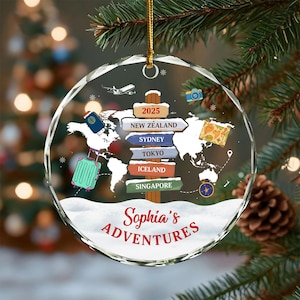 May include: A clear, round Christmas ornament with a world map design and the text "Sophia's Adventures." The ornament features a signpost pointing to New Zealand, Sydney, Tokyo, Iceland, and Singapore. A small airplane, camera, and suitcase are also depicted.