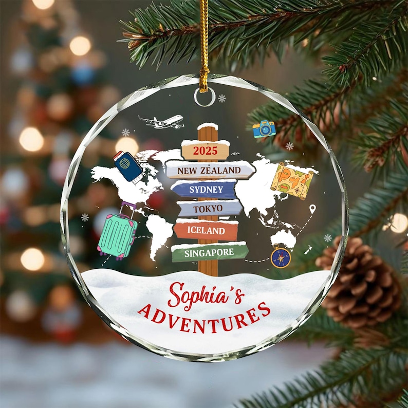 May include: A clear, round Christmas ornament with a world map design and the text "Sophia's Adventures." The ornament features a signpost pointing to New Zealand, Sydney, Tokyo, Iceland, and Singapore. A small airplane, camera, and suitcase are also depicted.