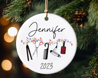 Op de afbeelding: Ronde witte keramische kerstversiering met de naam "Jennifer" in zwart script. Het heeft kappersgereedschap en kleurrijke lichtslingers. Het jaar "2023" staat onderaan gedrukt. Een bruin touwtje is bevestigd om op te hangen.