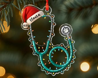 May include: A Christmas ornament shaped like a stethoscope, decorated with colorful lights. A red Santa hat with the name "Claura" and the year "2025" is attached. The ornament hangs from a red ribbon against a blurred background.