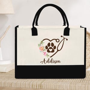 Peut inclure: Un sac fourre-tout en toile blanche avec des anses noires et un fond noir. Le sac présente un motif de cœur brun avec une empreinte de patte et un stéthoscope à l'intérieur du cœur. Le texte "Addison" est imprimé sous le cœur.