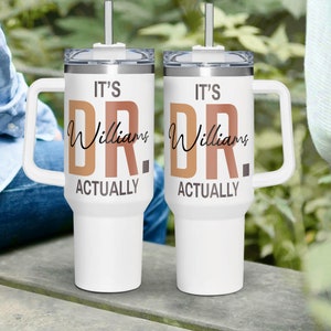 May include: Two white stainless steel travel mugs with handles and lids. The mugs have the text "It's Dr. Williams Actually" printed on them in brown and orange.