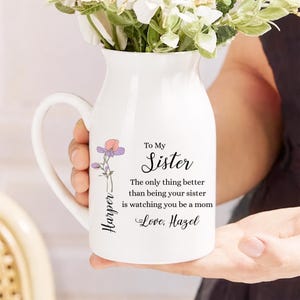 Peut inclure: Pichet en céramique blanche avec une anse, rempli de fleurs blanches et vertes. Le pichet porte une décoration florale et le texte "To My Sister" avec un message personnalisé. Un cadeau attentionné.