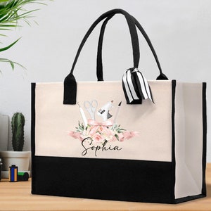 May include: A beige tote bag with black handles and a black bottom panel. The bag features a floral design with craft tools and the name "Sophia" in script. A black and white striped ribbon is tied to one handle.