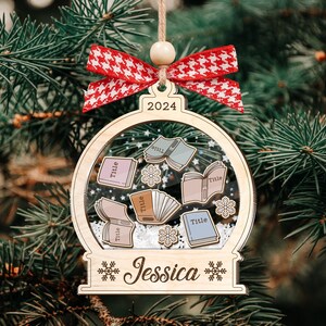 Op de afbeelding: Een houten kerstversiering in de vorm van een sneeuwbol met een witte achtergrond en een transparante koepel. De koepel bevat verschillende boeken met het woord "Titel" erop. De versiering heeft ook het jaar "2024" en de naam "Jessica" erop.