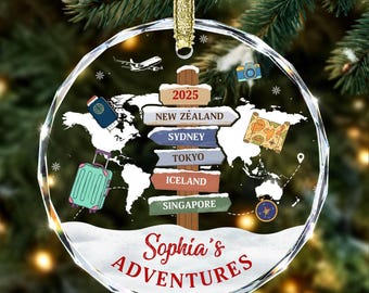 May include: A circular, faceted glass ornament featuring a world map background, a central wooden signpost with the text 2025, New Zealand, Sydney, Tokyo, Iceland, and Singapore, plus travel icons including a plane, camera, passport, map, and suitcase.