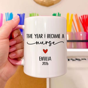 Op de afbeelding: Witte keramische mok met een roze binnenkant en handvat. De mok heeft de tekst "THE YEAR I BECAME A nurse" in zwart script, een rood hart en de naam "EMILIA" met het jaar "2026" eronder. De mok wordt door een hand vastgehouden.