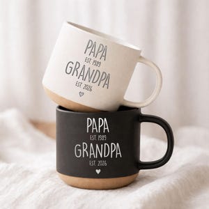 Puede incluir: Dos tazas de cerámica, una blanca y otra negra, apiladas. La taza blanca tiene una base beige y un asa blanca. La taza negra tiene una base beige y un asa negra. Ambas tazas tienen la inscripción "PAPA EST. 1989 GRANDPA EST. 2026".
