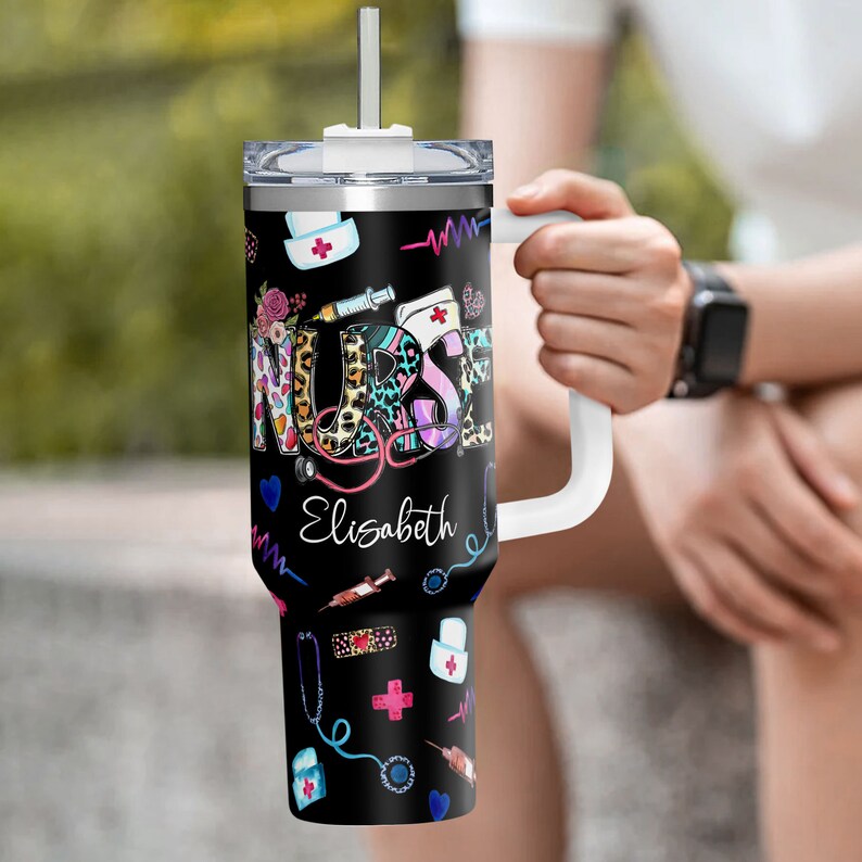 May include: A black and white stainless steel travel mug with a handle. The mug has a colorful design with the word "NURSE" and the name "Elizabeth". The design also includes medical symbols like a stethoscope, a syringe, and a bandage.