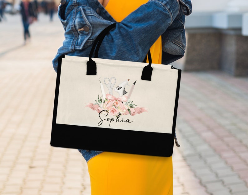 May include: A cream-colored tote bag with a black bottom and handles. The bag features a floral design with pink flowers, scissors, and a pen. The name "Sophia" is written in cursive below the floral arrangement.