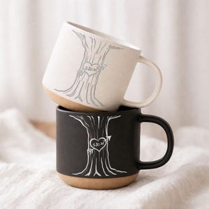 Peut inclure: Deux mugs en céramique, un crème et un noir, empilés. Chaque mug présente un motif d'arbre avec un cœur et des initiales. Les mugs ont une base de couleur naturelle et une anse incurvée. Les mugs sont probablement pour les boissons.