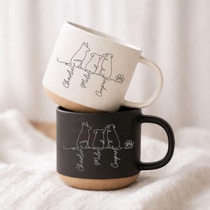 Könnte beinhalten: Zwei Keramikbecher, einer weiß und einer schwarz, gestapelt. Jeder Becher zeigt eine Strichzeichnung von drei Hunden und einem Pfotenabdruck, mit den Namen "Charlie", "Milo" und "Cooper" darunter. Die Becher haben einen natürlichen, hellbraunen Boden.