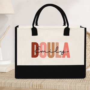 May include: A white canvas tote bag with black handles and a black bottom. The bag has the word "DOULA" printed in large, colorful letters with the name "Emily" written in cursive underneath.