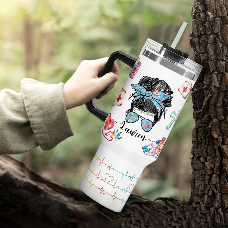 May include: White tumbler with a black handle and a straw. The tumbler features a design of a woman with a blue bandana and sunglasses, medical-themed illustrations, and the name "Lauren". A colorful heartbeat design is on the bottom.
