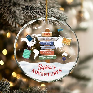May include: A clear, round Christmas ornament with a gold string. The ornament features a world map design with travel-themed icons and the text "Sophia's Adventures." Destinations include New Zealand, Sydney, Tokyo, Iceland, and Singapore.