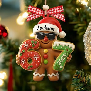 May include: A gingerbread man Christmas ornament wearing sunglasses and a Santa hat with the name "Jackson". The ornament holds the numbers "6" and "7" with the year "2025". A red and white checkered bow is at the top.