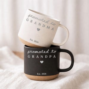 Peut inclure: Deux mugs en céramique, un blanc et un noir, empilés. Le mug blanc porte l'inscription "promoted to GRANDMA" avec un cœur et "Est. 2026". Le mug noir porte l'inscription "promoted to GRANDPA" avec un cœur et "Est. 2026".