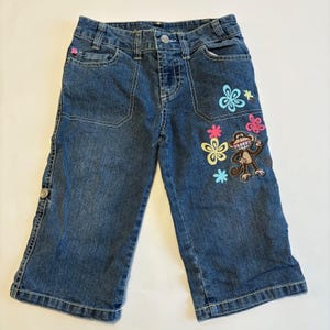 May include: Blue denim capri pants with a monkey and flower applique. The capri pants have two front pockets, a button and zipper closure, and decorative stitching. The monkey applique is brown with a smiling face, and the flowers are pink, yellow, and blue.