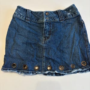 May include: A blue denim mini skirt with a distressed hem and metal grommets along the bottom edge. The skirt has a classic five-pocket design, a button and zipper closure, and belt loops. The denim is a medium wash.