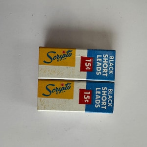 May include: Two vintage Scripto M.A. pencil lead boxes. The boxes are rectangular with a yellow top section featuring the Scripto logo in blue script. The bottom section is blue with the text "BLACK SHORT LEADS" and a red box with the price.