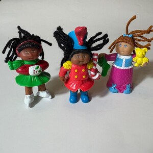 May include: Three Cabbage Patch Kids dolls. One doll wears a red and green outfit with a holly berry. Another doll is dressed as a toy soldier in a red uniform and blue hat. The third doll wears a pink dress and holds a yellow teddy bear.