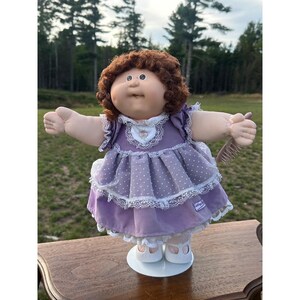 May include: A doll with curly brown hair and a round face wearing a lavender dress with white polka dots, lace trim, and a small floral accent at the neckline. The doll has white shoes and is holding a small brown plastic hairbrush in one hand.