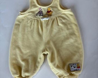 May include: A light yellow baby romper featuring embroidered images of Winnie the Pooh and Piglet. The romper has a scoop neckline, snap closures, and a small patch with text. The garment is made of a soft, textured fabric.