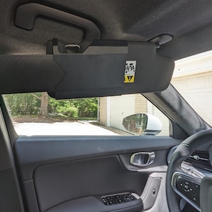 May include: A black sun visor with a yellow warning label attached to it. The visor is in a car interior.