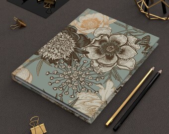 May include: A blue floral notebook with a vintage botanical design featuring peonies, leaves, and other flowers. The notebook is closed and lying on a textured surface.