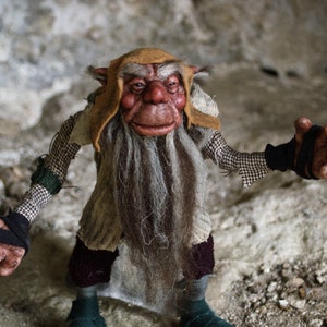 May include: A whimsical goblin puppet with a long white beard, wearing a brown and white hat and a black and white patterned shirt. The puppet has a mischievous expression and is holding out its hand.