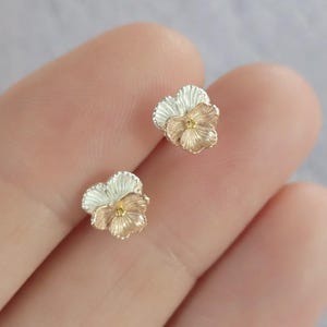 Puede incluir: Un par de pendientes de botón de flor de plata y oro. Las flores son de color rosa claro y blanco con un centro dorado.