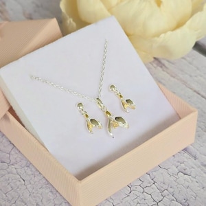 May include: A silver necklace and earrings set featuring a delicate snowdrop flower design with gold accents. The jewellery is displayed in a pink box.