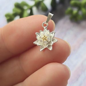 Puede incluir: Un colgante de plata en forma de flor de loto blanca con un centro dorado. El colgante está colgado de una cadena de plata.