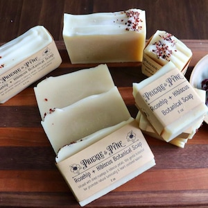 May include: A selection of Rosehip + Hibiscus Botanical Soap bars, light beige with red petal decoration, are arranged on a wooden surface. The packaging features the brand "Prickle & Pyne". A small plate holds dried hibiscus flowers. The soap bars are approximately 8 cm long.