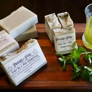 May include: A wooden cutting board displays several bars of green tea and mint infused soap, labeled "PRICKLE & PYNE". A glass of green liquid and fresh mint sprigs are also present. The soap bars are light green with visible herbs.