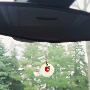 May include: A red apple with a yellow heart charm hanging from a rearview mirror in a car. The apple is on a gold-colored round pendant.