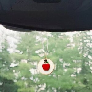 May include: A red apple charm hanging from a white string, with a green leaf on top. The charm is round and has a white background. The apple is hanging from a car's rearview mirror.