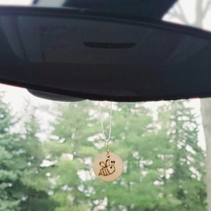 May include: A round, wooden car air freshener with a laser-etched bee design. The charm is suspended by a thin, white string and hangs from a car's rearview mirror. The background shows a blurred view of green trees.