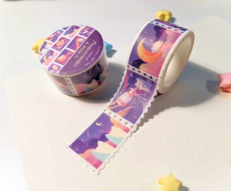 Pu&ograve; includere: Un rotolo di nastro washi viola e bianco con un motivo ripetuto di stelle, lune e pianeti. Il nastro ha la scritta "a little astronomy" stampata su di esso.
