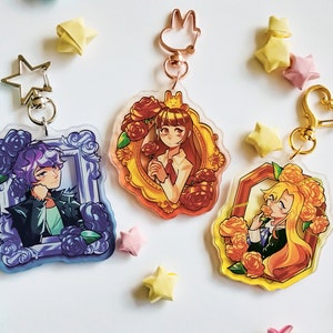 Può includere: Tre portachiavi con personaggi colorati e anelli in metallo dorato. Ogni portachiavi presenta un personaggio diverso con un design floreale. I personaggi sono incorniciati in una cornice dorata, argentata e rossa.