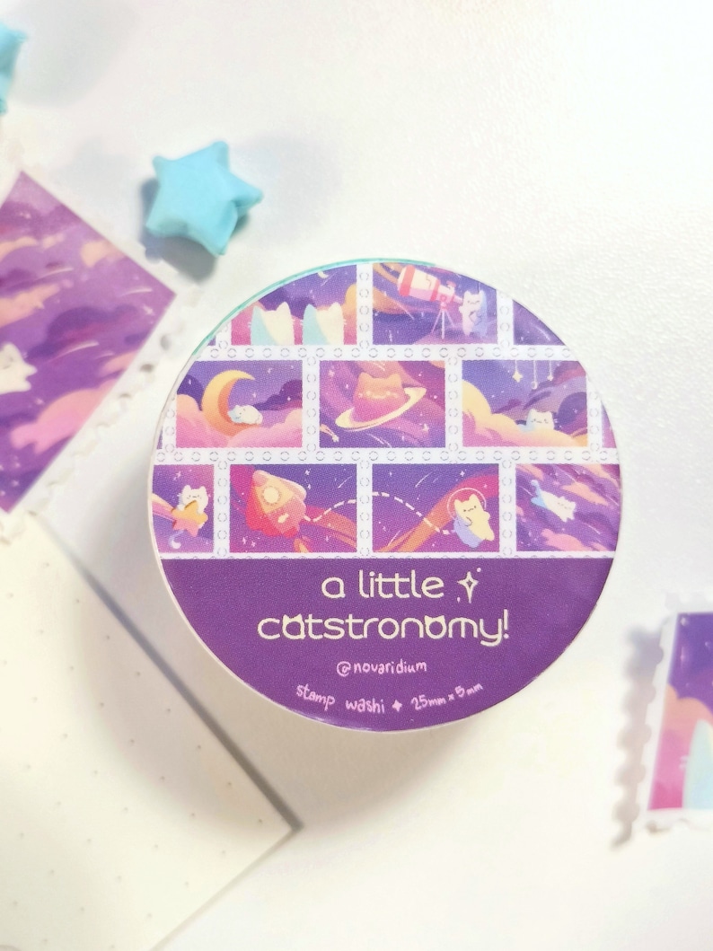 Pu&ograve; includere: Un rotolo di nastro washi viola e blu con un motivo ripetuto di gatti nello spazio. Il nastro &egrave; etichettato "a little catstronomy!" e "@novaridium stamp washi + 25mm x 8mm".