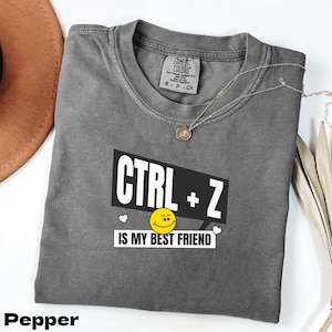 Op de afbeelding: Een grijs t-shirt met de tekst "CTRL + Z IS MY BEST FRIEND" in wit en zwart. Het shirt heeft een smiley-gezicht en een kleine hartafbeelding. Een zilveren ketting met een hanger ligt op het shirt.