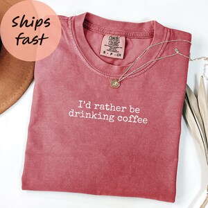 May include: A dusty rose-colored t-shirt with the text "I'd rather be drinking coffee" in white. A silver necklace with a round pendant rests on the shirt. The shirt is folded and displayed on a white surface.