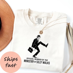 May include: Off-white t-shirt with a black graphic of a man in a bowler hat and suit, walking with a raised leg, holding a briefcase. The text below reads "OFFICIAL MEMBER OF THE MINISTRY OF SILLY WALKS". A gold necklace and a brown hat are also visible.