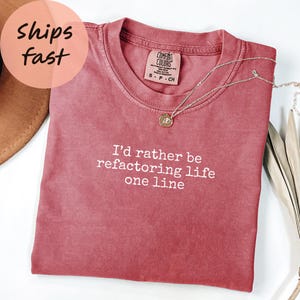 以下が含まれることがあります： 「I'd rather be refactoring life one line」の白文字が入った、くすんだバラ色のComfort Colors Tシャツ。丸いペンダントが付いたシルバーのネックレスがシャツの上にあります。シャツは白い表面に折りたたまれています。