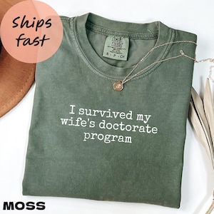 May include: A moss green t-shirt with the text "I survived my wife's doctorate program" in white. A gold necklace with a pendant rests on the shirt. The shirt is labeled "Comfort Colors" and is size small.