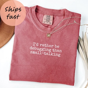 Puede incluir: Una camiseta rosa descolorida con el texto blanco "I'd rather be debugging than small-talking". Un collar con colgante dorado descansa sobre la camiseta. La camiseta es de Comfort Colors.
