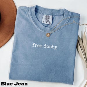 以下が含まれることがあります： 「free dobby」の白い文字が書かれた、ブルーデニムのComfort Colors Tシャツ。 金色のネックレスとペンダントがシャツの上にあります。 茶色の帽子と乾燥した葉が背景にあります。